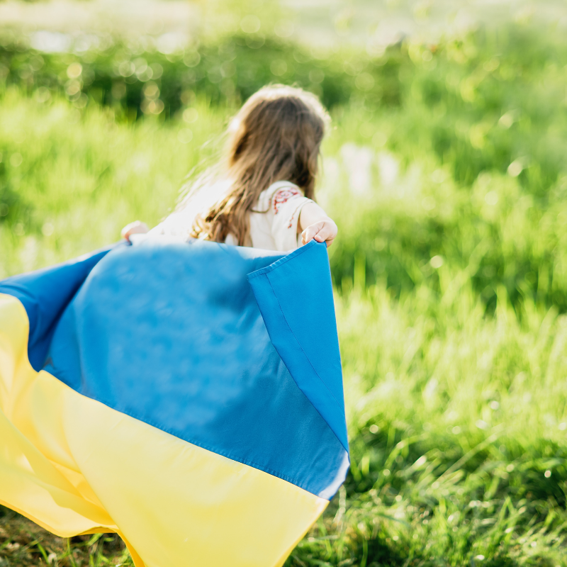 Dziewczynka z flagą Ukrainy biegnąca po trawie. Na dole malinowa apla: Co dzieci powinny wiedzieć o wojnie w Ukrainie.
